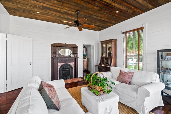 Spacious living room with white shiplap walls, exposed wooden beam ceiling, fireplace with ornate mantel, white upholstered seating, and glass doors opening to