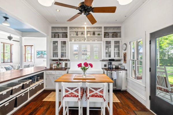 Bright kitchen with white cabinetry, wood island with pink flowers, ceiling fan, and glass doors opening to green lawn.