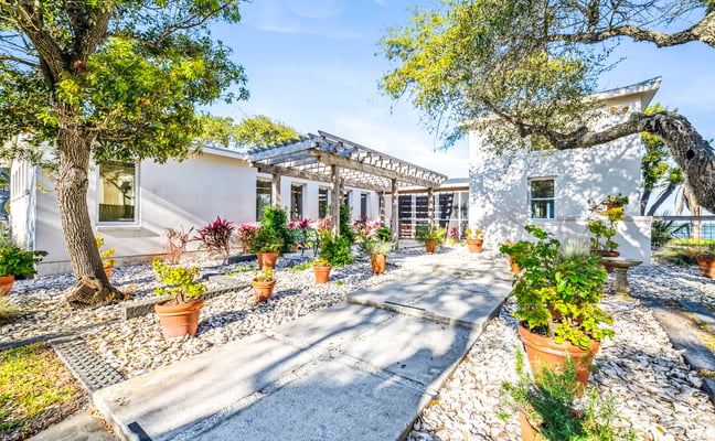Modern white house with pergola, gravel courtyard, potted flowers, and mature trees providing shade